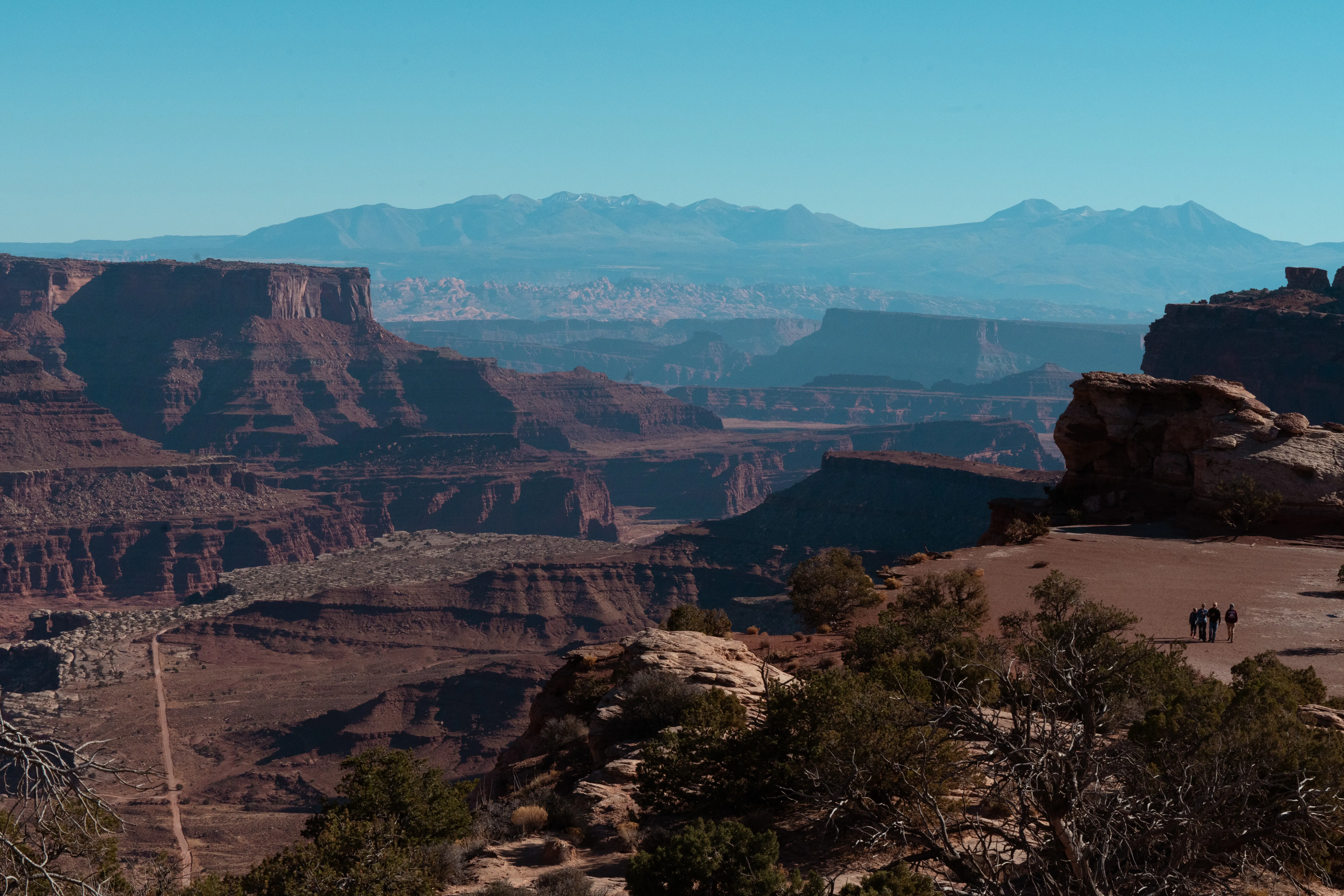 Layered red-rock canyons and mesas at a Canyonlands overlook with distant mountains beyond and tiny figures at the rim