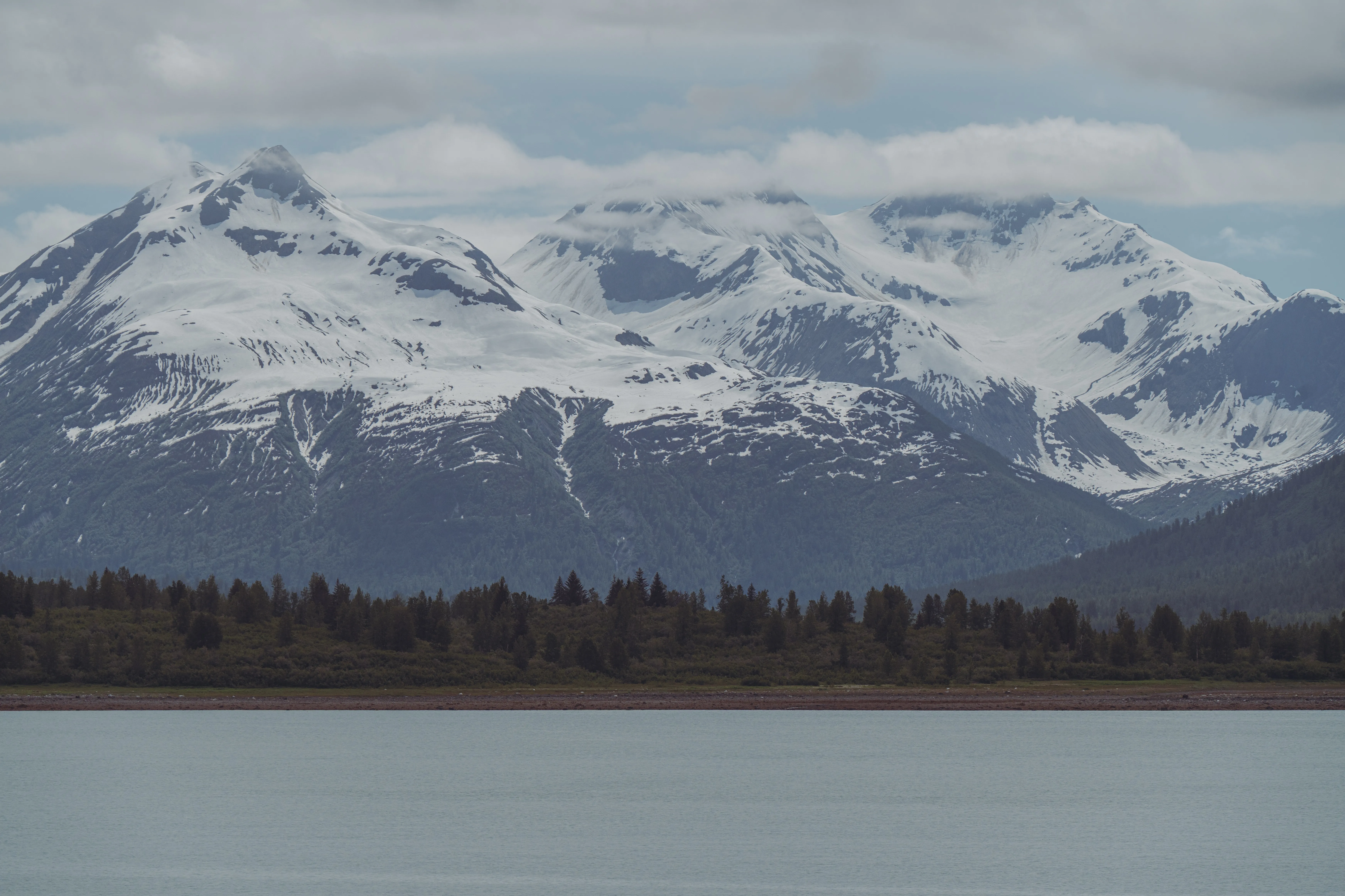 Cloud-draped snow-capped mountains rising above a forested shoreline and calm water