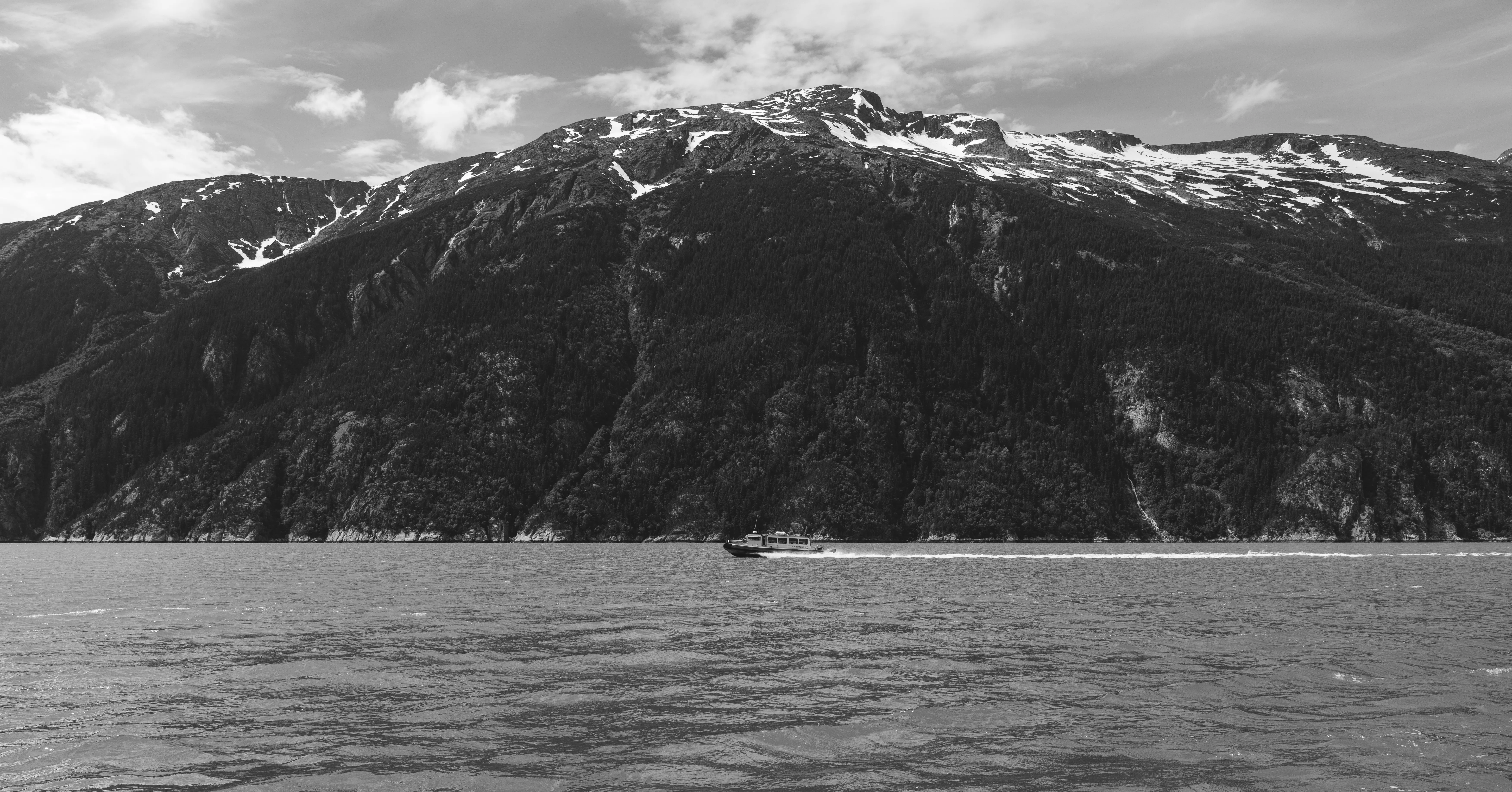 Black-and-white view of a forested mountain rising from the water with a small boat crossing below