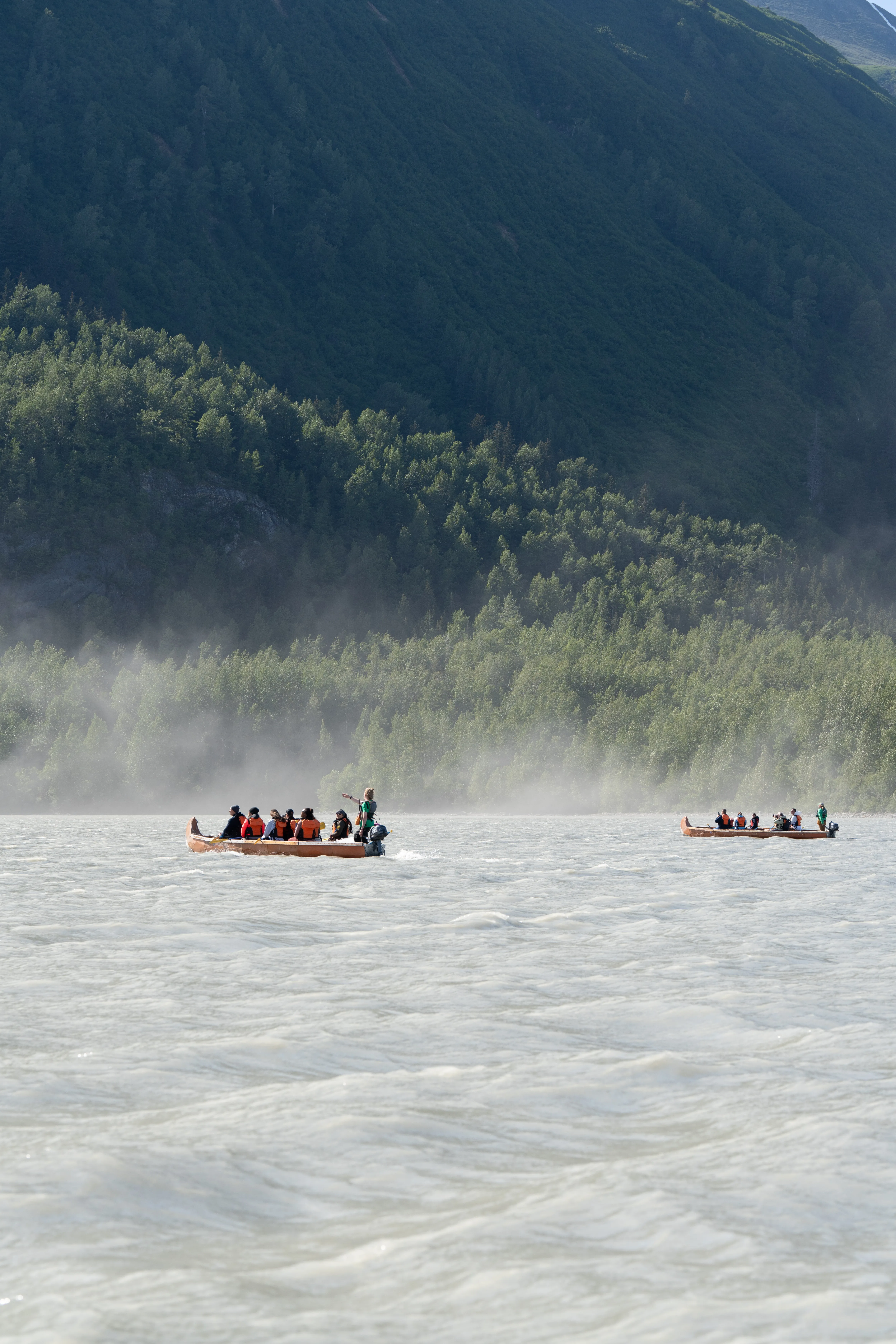 Two canoes of paddlers crossing a misty, silt-laden glacial river below a forested slope