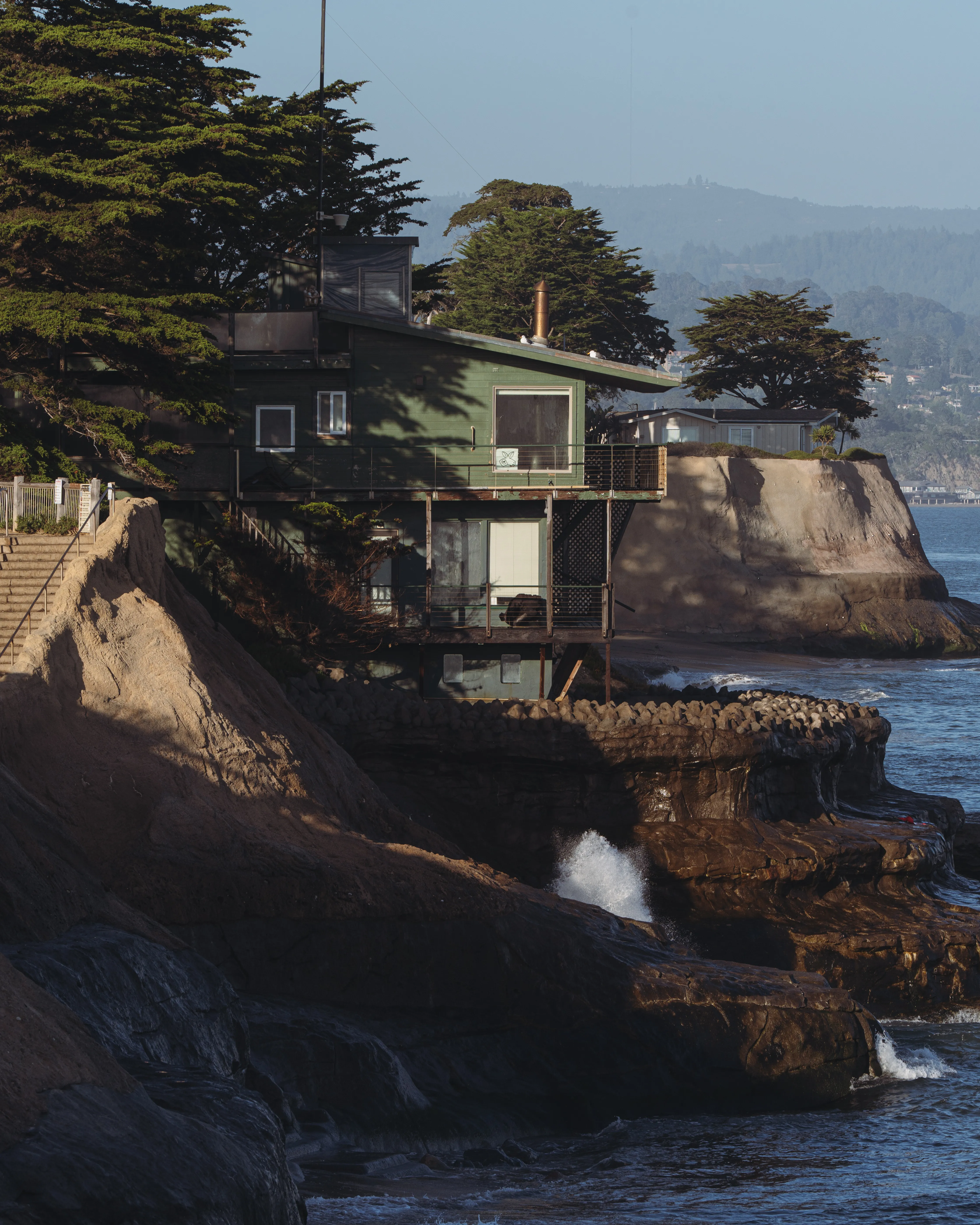 A green clifftop house perched on eroding coastal bluffs above the ocean at golden hour