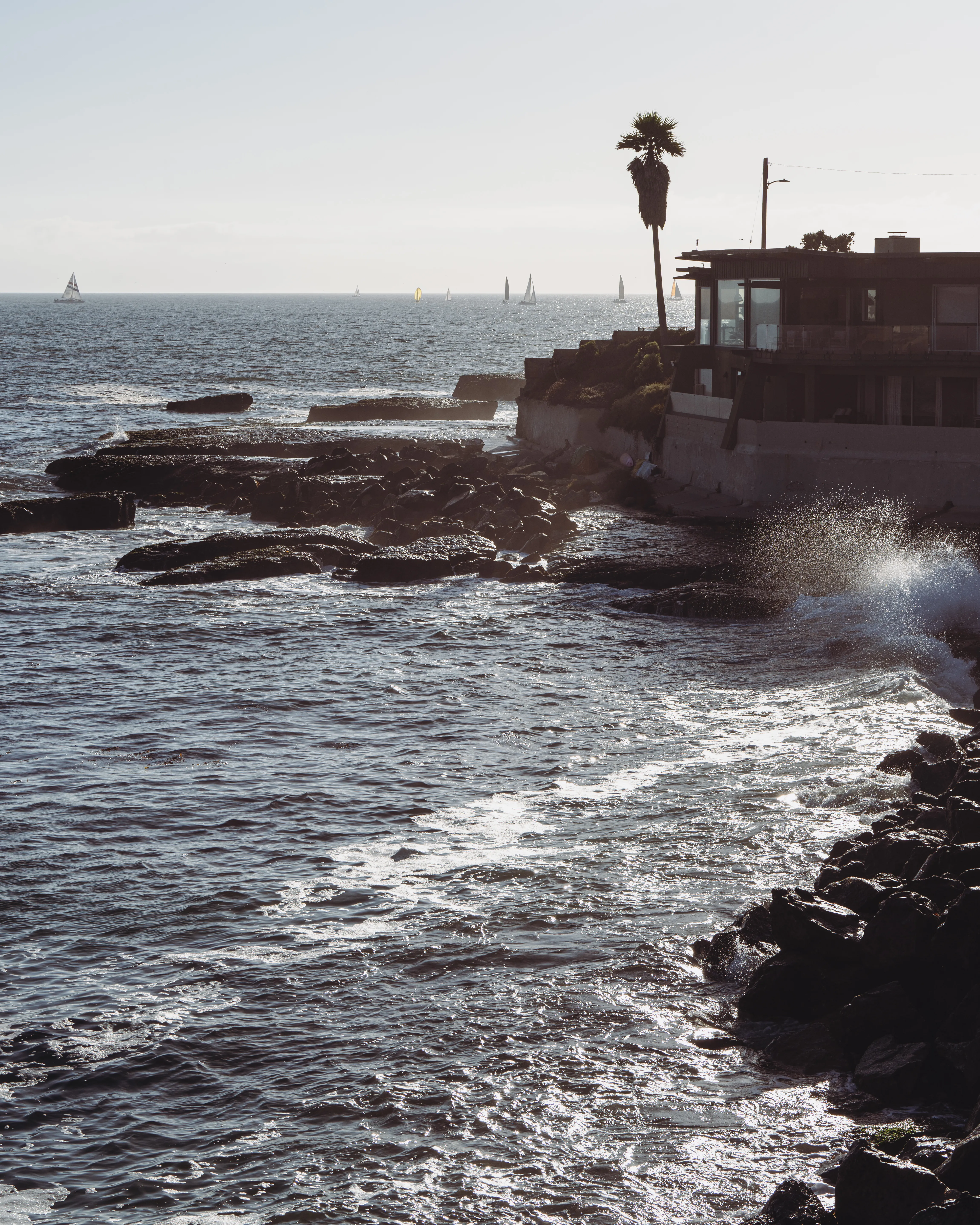 Waves breaking over a rocky shoreline at golden hour with sailboats offshore and a clifftop house on the point