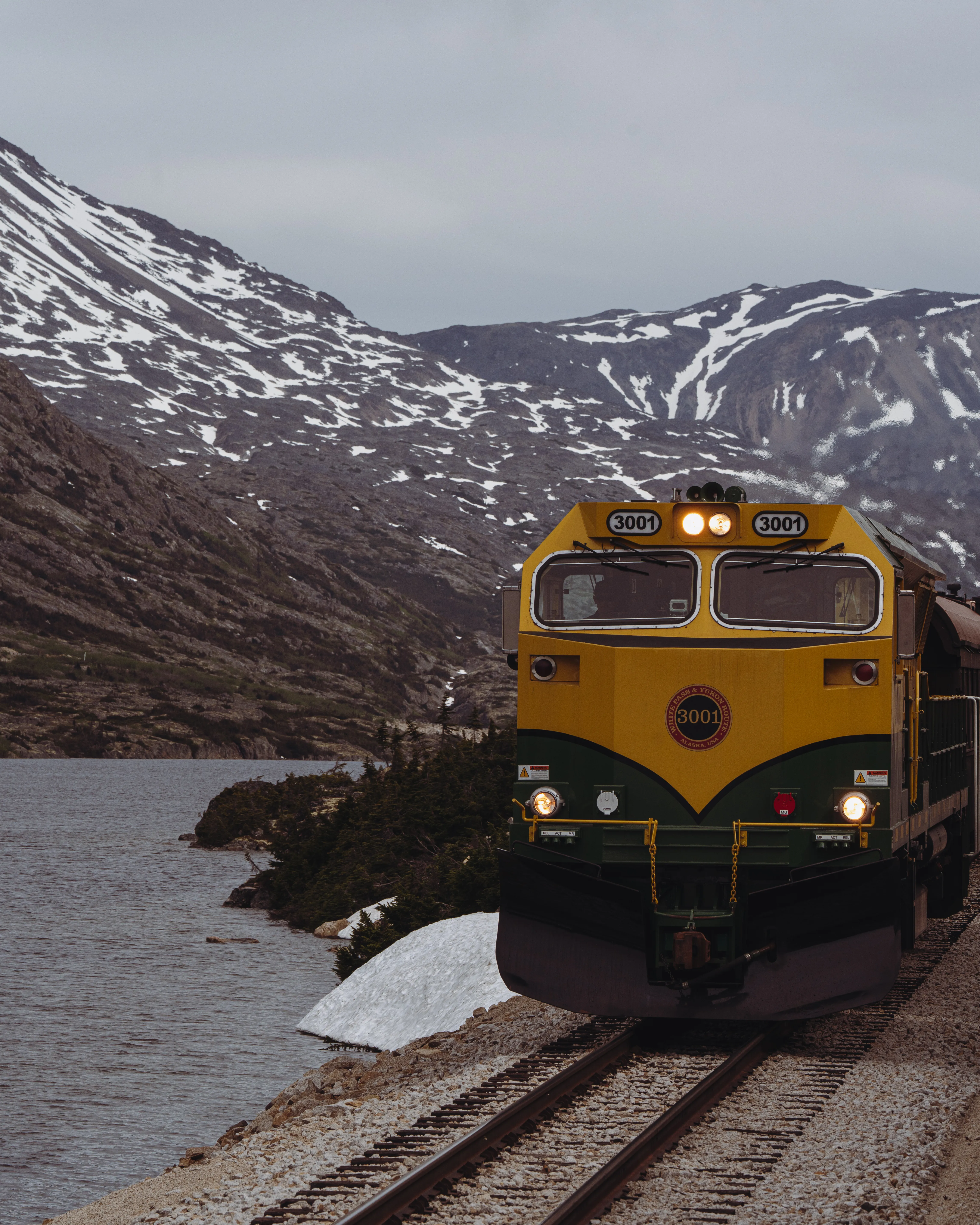 The yellow-and-green White Pass locomotive on the tracks beside a lake below snow-patched mountains